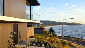 Ein modernes Küstengebäude mit Balkon und Meerblick, eine Straßenlaterne sowie ein freier Blick auf den Strand und die Hügel im Hintergrund.