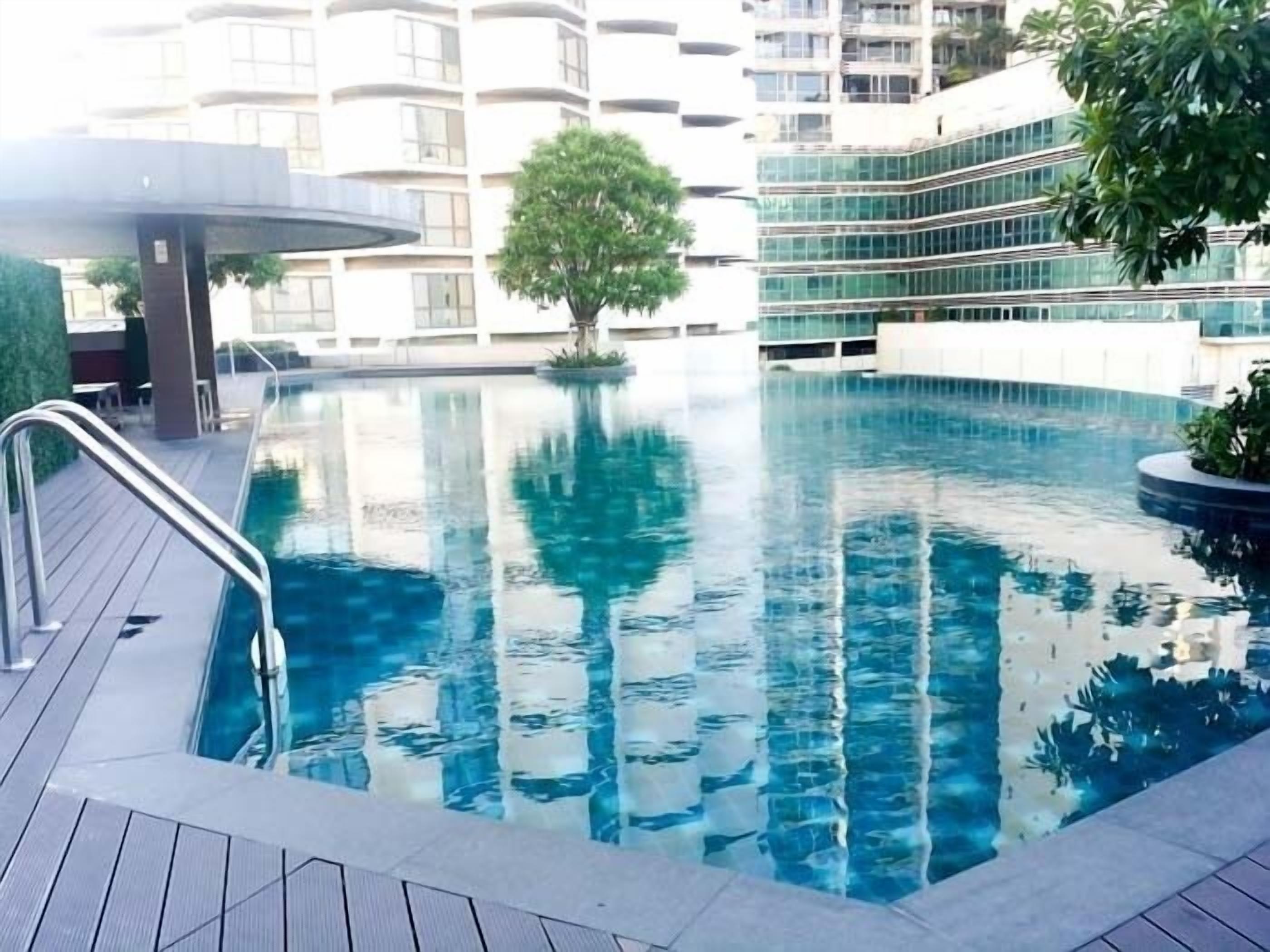 Une piscine dotée d’une terrasse en bois, entourée de bâtiments modernes et d’un arbre.