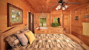 Ein Schlafzimmer in einer Holzhütte mit einem Bett, einem Schminktisch, einem Deckenventilator und einem Fenster mit Blick auf Grünflächen.