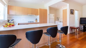 Una cocina moderna con armarios de madera, una encimera blanca y un frutero. Hay tres taburetes negros y una pequeña mesa de comedor con un televisor.