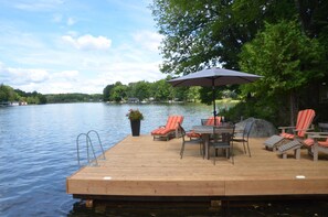 Un muelle de madera con sillones de descanso y una mesa, con vista a un lago tranquilo con árboles y casas al fondo.