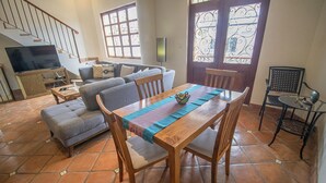 Een woonkamer met een bank, een houten eettafel en stoelen. Er is een televisie op een dressoir, een raam met sierlijk ijzerwerk en een deur met ruit.