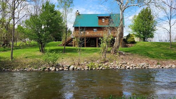 Eine Blockhütte mit grüner Dachdeckung, einer Veranda und einem Fluss im Vordergrund.