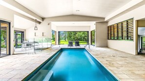 A modern indoor swimming pool with clear glass walls, two seating areas, and a view of a green landscape.