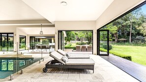 A modern outdoor lounge area with a glass-enclosed pool, a sun lounger, and a dining area with outdoor furniture.