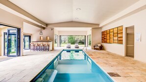 Un espace piscine intérieur moderne, doté d’une longue piscine, d’un bar avec des sièges et d’une vue sur l’extérieur.