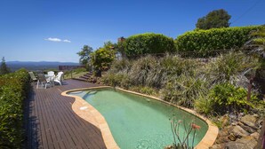 A swimming pool with a wooden deck, surrounded by various plants and rocks.