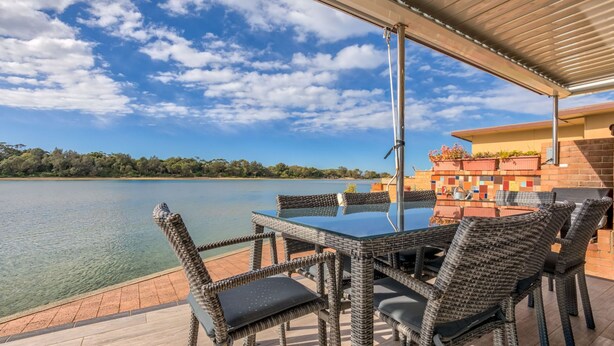 A covered outdoor dining area with wicker furniture and a glass table, overlooking a calm lake and distant trees.
