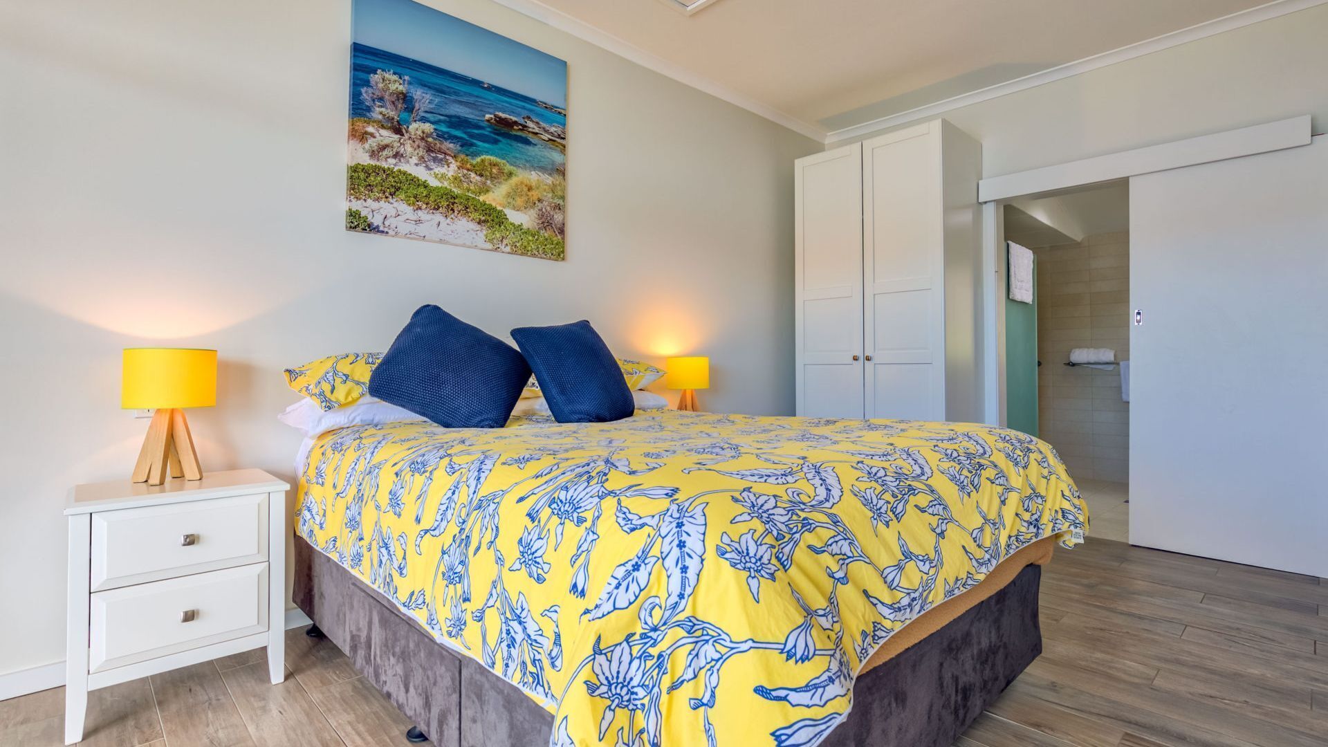 A bedroom with a bed, two bedside lamps, a nightstand, a wardrobe, and a painting of a beach scene.