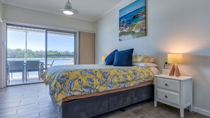 A bedroom with a bed, a nightstand, a lamp, a painting, and a view of the outdoors.