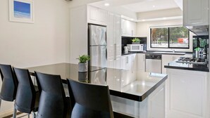 Una cocina moderna con armarios blancos, un refrigerador de acero inoxidable, un microondas y una estufa a gas. Hay una mesa de comedor con sillas y una planta en maceta.