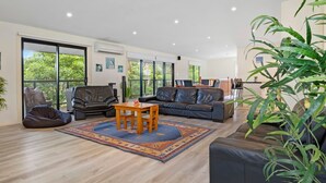Amplia sala de estar con un sofá de cuero, una mesa de centro de madera y un sillón informal. Amplias ventanas ofrecen vistas a la vegetación exterior.