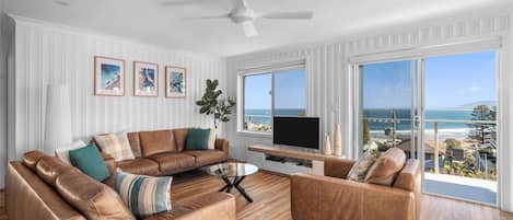 Een moderne woonkamer met een groot raam met uitzicht op de oceaan, een plafondventilator en een flatscreen televisie op een houten console.