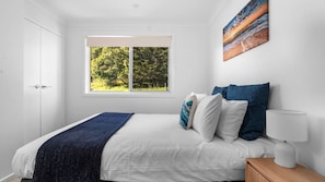 A bedroom with a bed, pillows, a bedside table, a lamp, and a window with a view of trees.