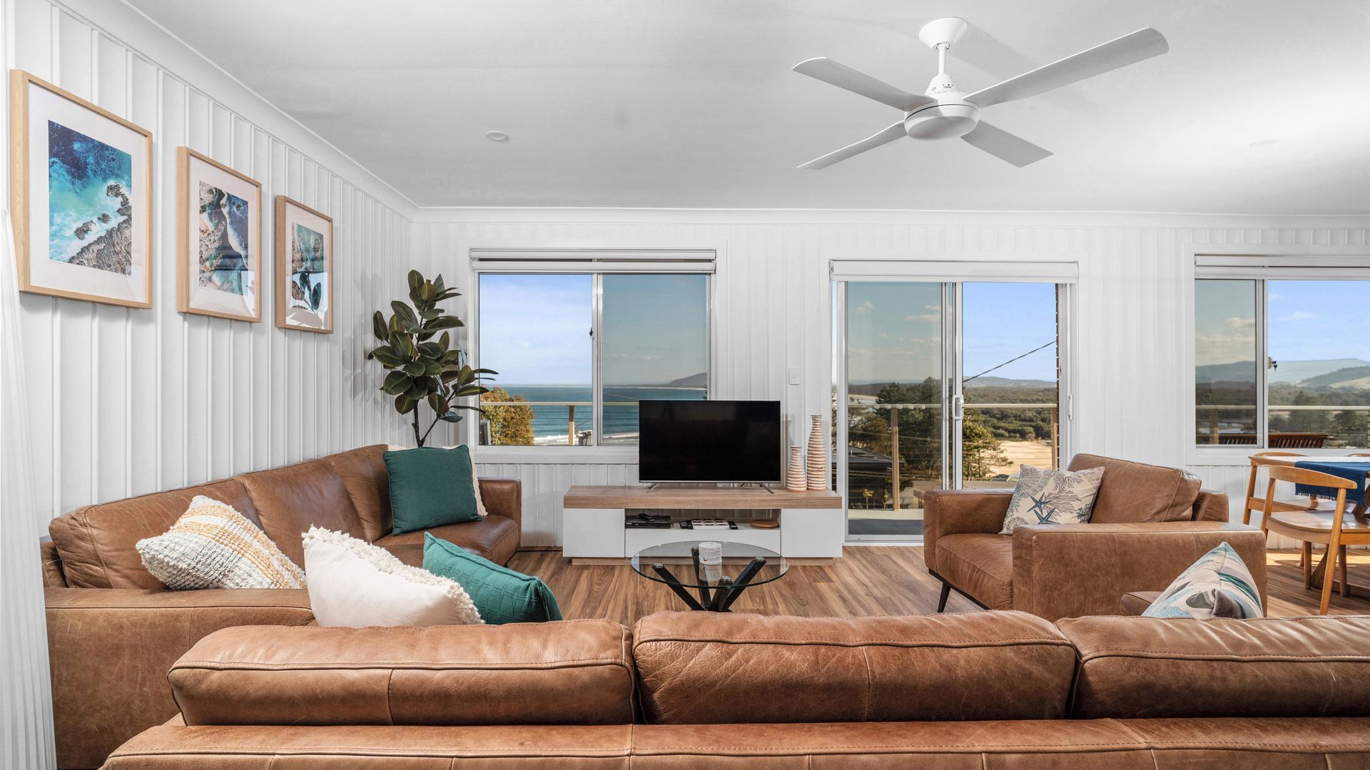 A spacious living room with a brown leather sofa, a flat-screen TV, and a ceiling fan.