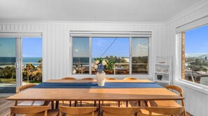 A dining area with a wooden table and chairs, a vase with flowers, and large windows with a view of the sea.