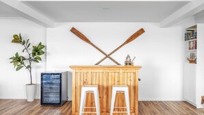 A room with a wooden bar area, two white stools, a potted plant, and a bookshelf. - Sunset on Headland, Gerroa (Gerroa)