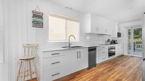 A modern kitchen with white cabinetry, a wooden stool, and a large window. - Sunsets on Headland, Gerroa (Gerroa)