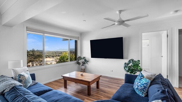 A modern living room with a large window, a flat-screen TV, a ceiling fan, and a wooden coffee table. - Sunsets on Headland, Gerroa (Gerroa)