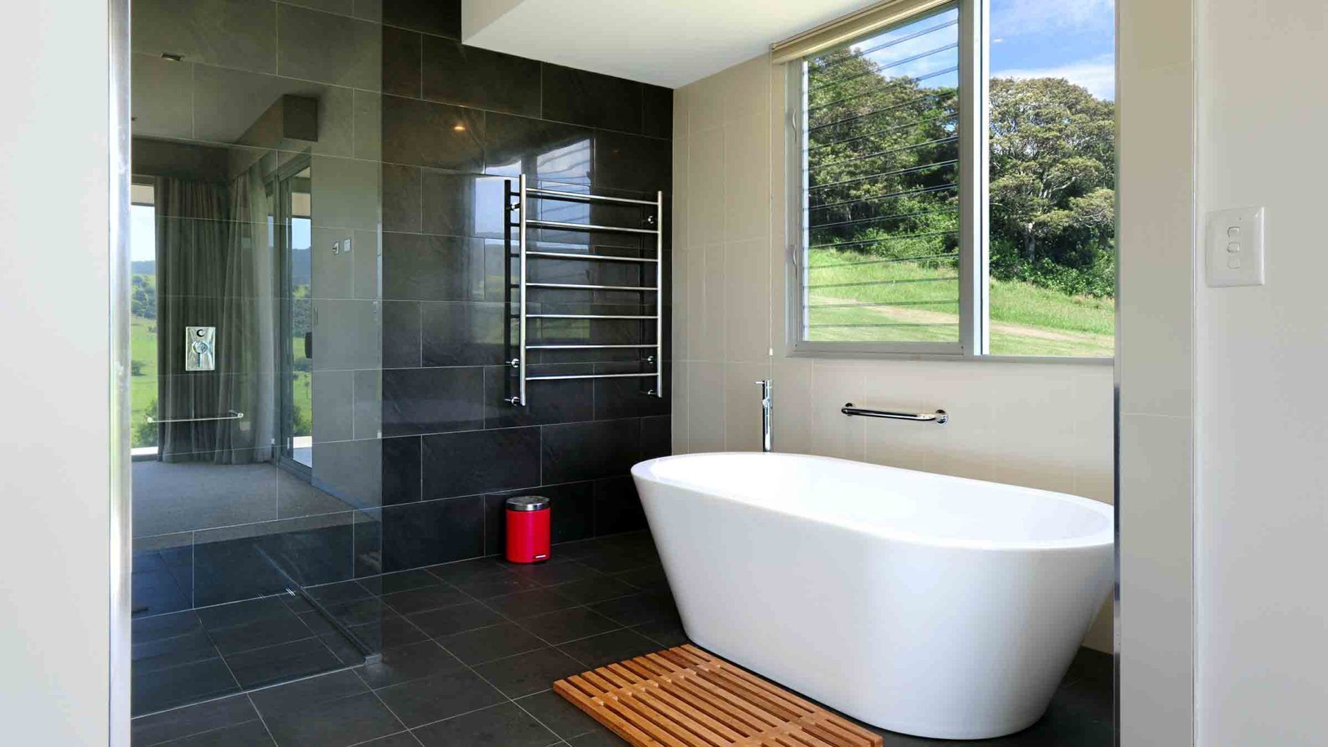 A modern bathroom with a freestanding bathtub, a glass shower enclosure, and a view of a green landscape through a large window.