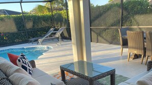 Zona d'estar a l'aire lliure coberta, amb mobiliari de vimet, taula de vidre i una piscina al fons.