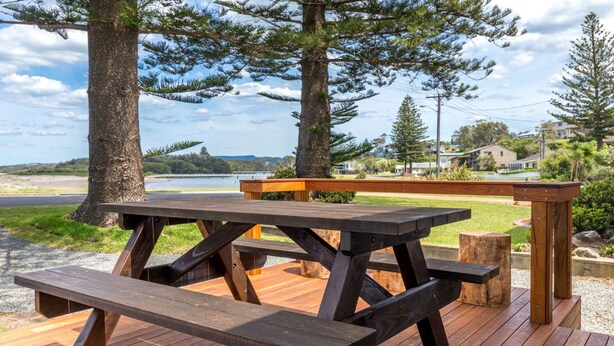 A wooden picnic table with benches, surrounded by trees and a view of a lake and buildings in the background. - Seaways, Gerroa, South Coast (Gerroa)