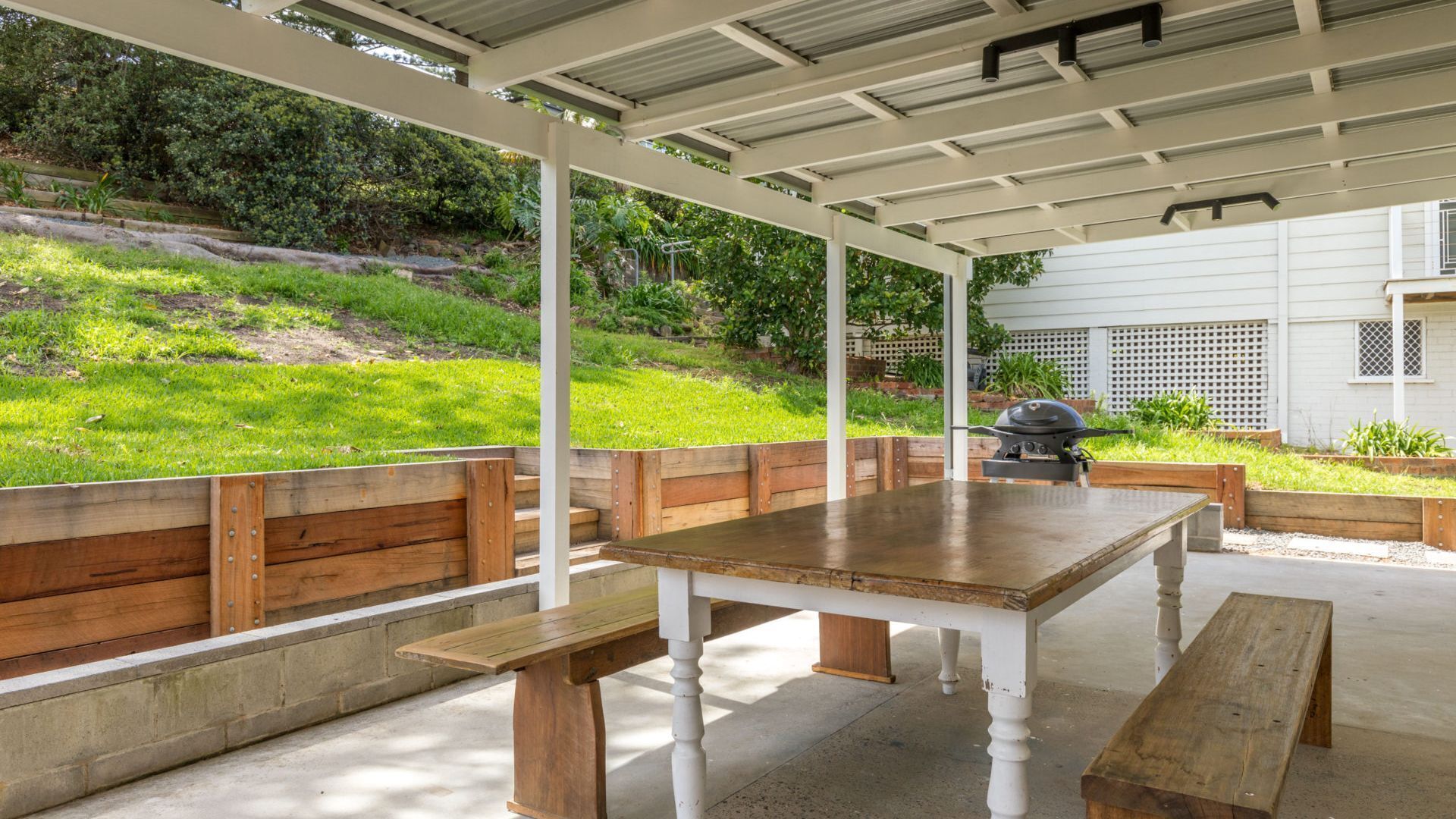 An outdoor patio with a wooden table and benches, surrounded by a white pergola.