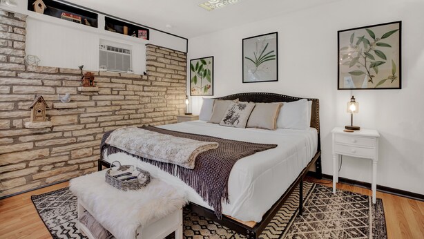A bedroom with a stone wall, a bed with a wooden frame, a white bedside table, and a patterned rug.