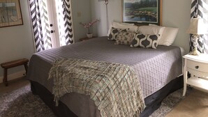 Quarto bem arrumado com uma cama grande, uma mesinha de cabeceira com abajur, um quadro na parede e um pequeno banco.