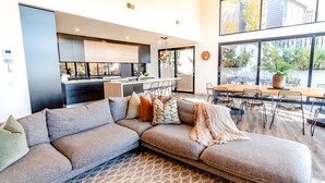 A modern living room with a sectional sofa, a dining area with a wooden table and chairs, a kitchen with dark cabinetry, and large windows offering a view of the outdoors. - GLASS HOUSE: Bright Township (Bright)