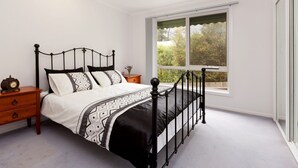 Quarto com cama, mesa de cabeceira, cômoda e janela com vista para vegetação.