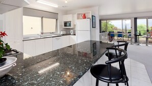 Cozinha moderna com bancada de granito, armários brancos, micro-ondas e geladeira. Há duas cadeiras pretas e uma mesa de jantar com cadeiras na parte externa.