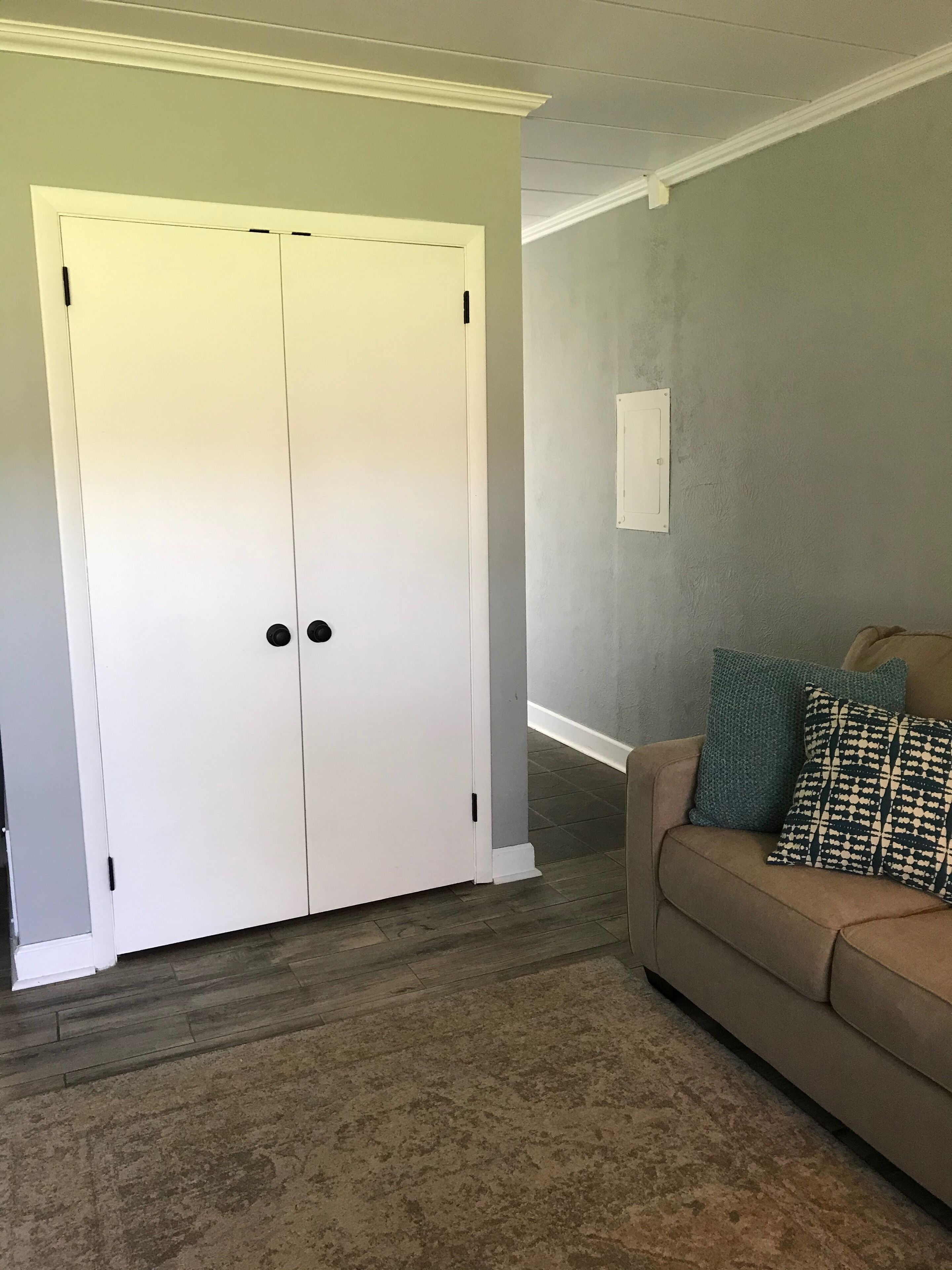 Ein Zimmer mit einem weißen, geschlossenen Kleiderschrank, einem braunen Sofa mit gemusterten Kissen und einer grauen Wand.