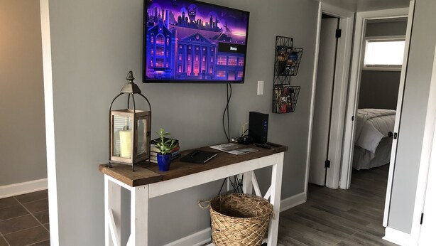 A modern living room with a TV mounted on the wall, a wooden console table, a floor lamp, a potted plant, and a basket. - Capital City Condo #1 close to Interstate, Water Park, Restaurants and Shopping (Frankfort)