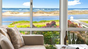 A room with a large window overlooking a beach and ocean, a sofa, and a table with a bottle and glasses. - The Beach House, Gerroa, South Coast (Gerroa)
