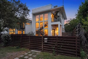 A modern two-story house with large windows, a visible staircase, and a wooden fence.