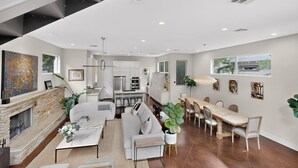 A modern living room with a fireplace, a dining area with a long table, and a kitchen in the background.