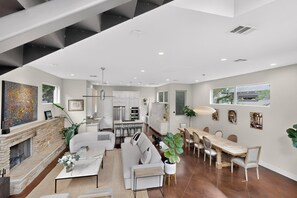 A modern living room with a fireplace, a dining area with a long table, and a kitchen in the background.