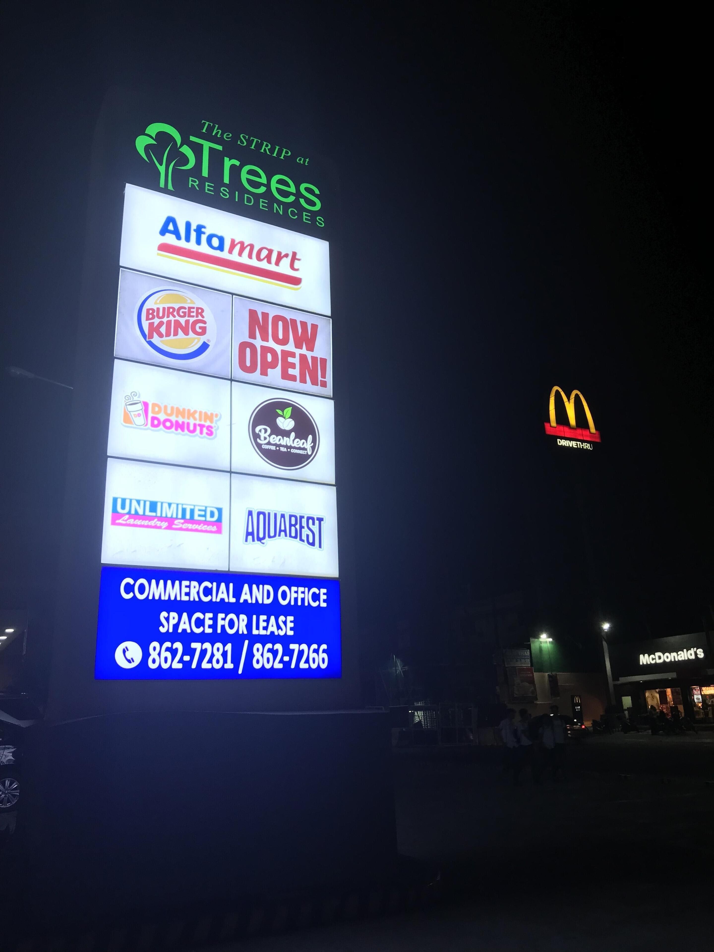 Un cartel nocturno publicita diversos negocios, incluyendo Almar Mart, Burger King, Dunkin' Donuts, Beanleaf Coffee, Servicios de Lavandería Ilimitados, Aquabest y McDonald's.