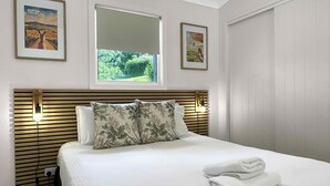 A neatly made bed with a white duvet, two pillows with a floral pattern, and a folded towel on the right side. There are two framed pictures on the wall, one depicting a vineyard and the other a landscape. - Villa 3 @ Bimbadeen Estate - Split level villa with views that stretch on forever - just for 2! (Mount View)