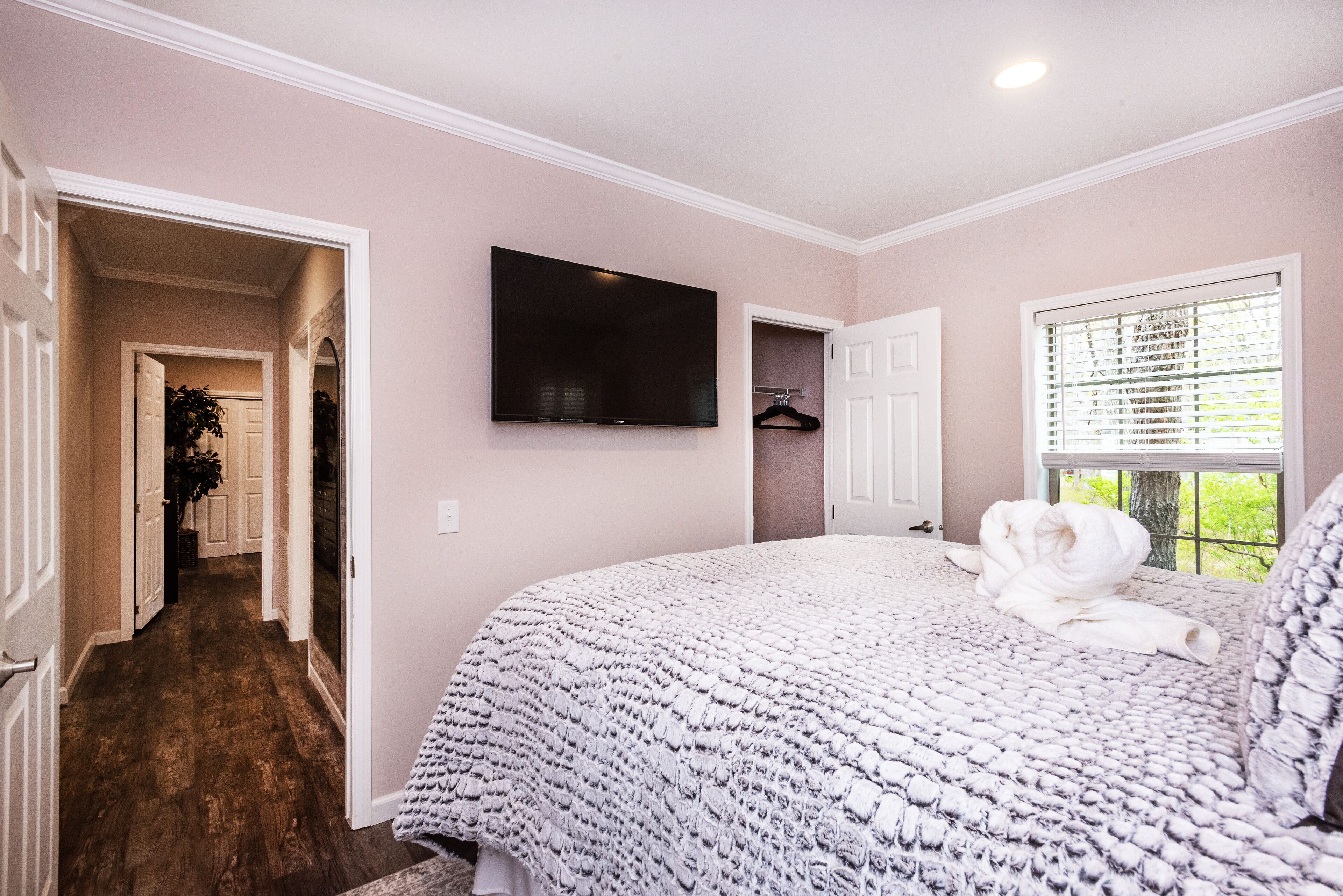 A bedroom with a large bed, a TV mounted on the wall, a window with blinds, and a hallway leading to another room.