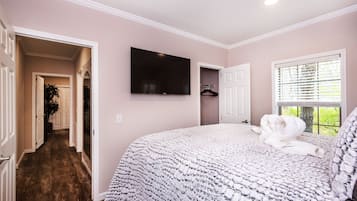 A bedroom with a large bed, a TV mounted on the wall, a window with blinds, and a hallway leading to another room.
