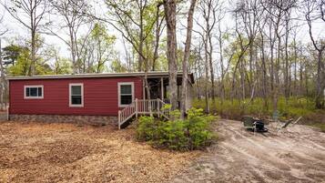 A red cabin with a wooden deck and a dirt path leading to it, surrounded by trees and shrubs.