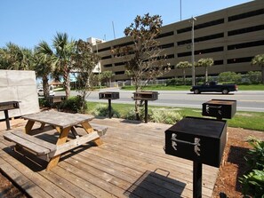 A wooden deck with picnic tables and a barbecue grill, surrounded by palm trees and a parking garage in the background. - *Wonderful Views!* Gulf Front! Aug 15 - 19th open! (Panama City Beach)