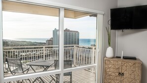 Un balcón con vista al océano y edificios, un televisor montado en la pared, y un mueble de madera con cajones.