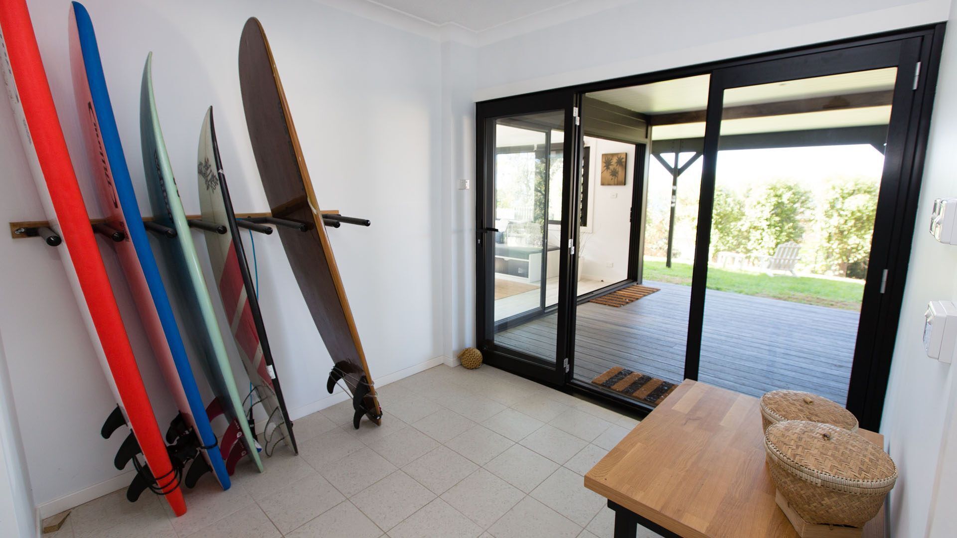 A room with a sliding glass door leading to a deck, a wooden table, and several surfboards leaning against the wall.