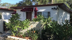 A single-story house with a red awning, surrounded by various plants and a clear blue sky.