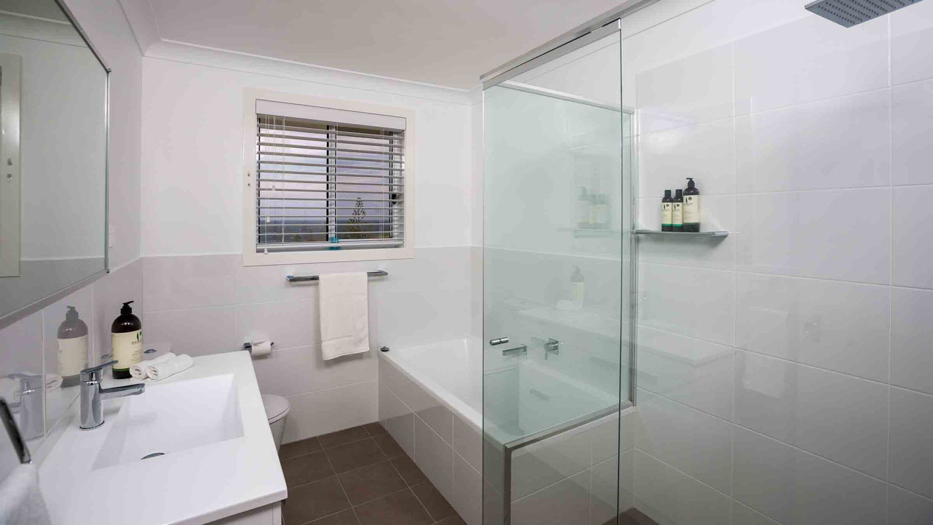 Un baño moderno con cabina de ducha de vidrio, un lavabo blanco y una ventana con persianas.
