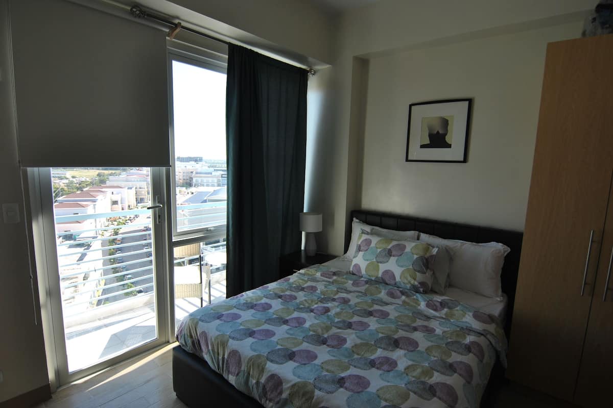 Quarto com cama, mesa de cabeceira, abajur, quadro na parede e vista através de uma porta de correr.