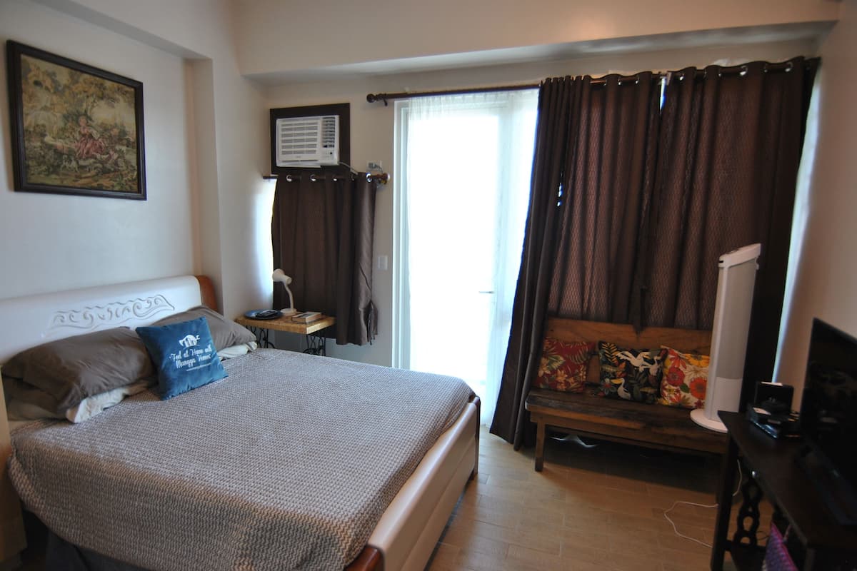 Quarto com cama, cadeira, escrivaninha, televisão e janela com cortinas.